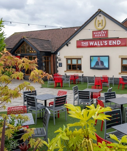 The exterior facade, signage, and beer garden seating area at The Wall's End, with string lights above the tables and chairs, and a TV on the wall.