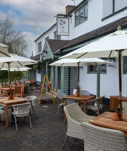 The exterior facade, signage, and seating area of The Hollybush, with shade umbrellas on some of the wooden tables, and flower baskets hanging from the veranda.