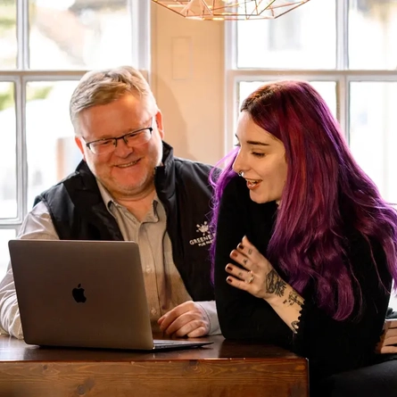 A Man and a Woman sat at a table looking at a laptop and smiling.