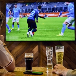 Two people sitting at a table in a pub, watching sports on the TV on the wall in front of them. A glass of Guinness, two glasses of Peroni, and a mobile phone sit on the table.