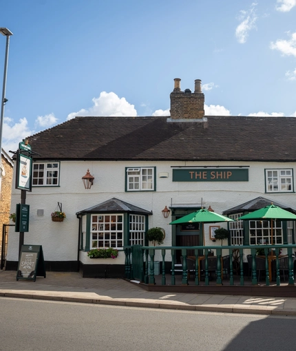 A view from across the road of the exterior facade, signage, and seating area of The Ship in Bedford; with shade umbrellas on some of the tables, and flower baskets and golden lanterns hanging on the white walls of the pub.