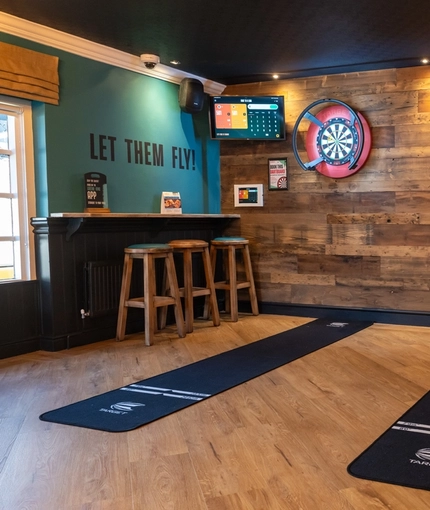The interior games area at The Lodge in Alvaston, with two dartboards, TVs on the wall, and bar stool seating.