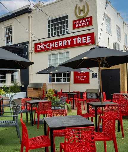 The exterior facade, signage, and beer garden seating area at The Cherry Tree; with shade umbrellas on some of the tables, and string lights overhead.