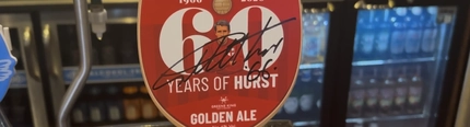 A close up image of a 60 Years of Hurst Golden Ale pump clip sat on an interior bar.