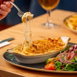A hand holding a fork taking a piece of macaroni cheese from a plated roasting dish on a wooden table.