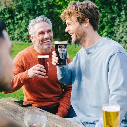 Four people at a wooden table in a sunny beer garden, two of them raising their glasses of Belhaven Best and Belhaven Black.