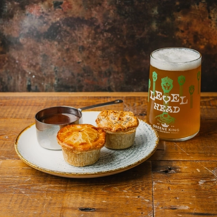 An advertising image showing 2 pies and a jug of gravy on a plate sat on a wooden table next to a pint of Level Head to advertise a Pie & Pint offer available at Urban Social & Community Social venues.