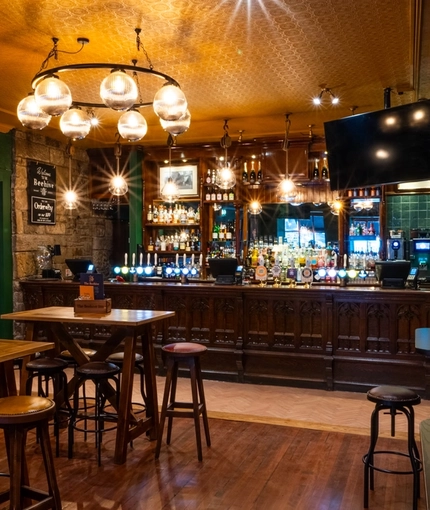 The bar and interior restaurant seating area at The Beehive Inn, with a TV hanging from the ceiling, bar stool seating, and gothic window style carvings on the front of the bar.