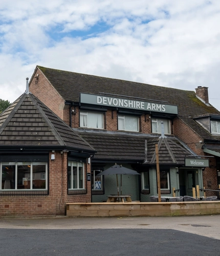 The exterior facade, signage, and seating area at the Devonshire Arms in Sheffield, with an accessibility ramp, wooden picnic tables, shade umbrellas, and string lights.