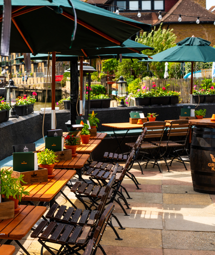 The exterior beer garden seating area which overlooks the River Thames, at The London Apprentice in Isleworth; with wooden tables and chairs, padded bench seats, shade umbrellas, and planter boxes filled with flowers on the stone wall enclosing the seating area.