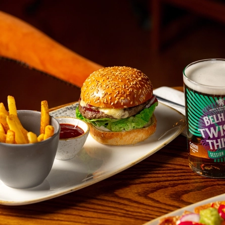 A close up view of a burger, a small bowl of fries, and a pot of sauce served on a plate which sits on a wooden restaurant table along with a glass of Belhaven Twisted Thistle IPA.