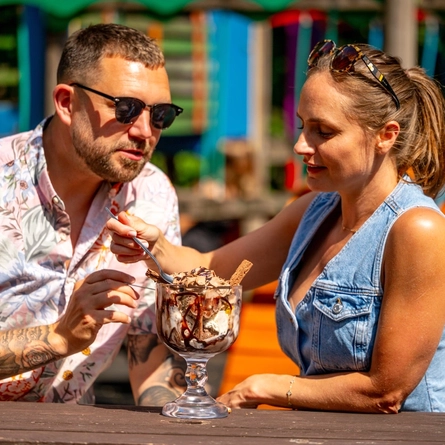 A couple sharing an ice cream sundae sharer whilst sat at an outdoor table.  There is the blurred background showing a child's play area.