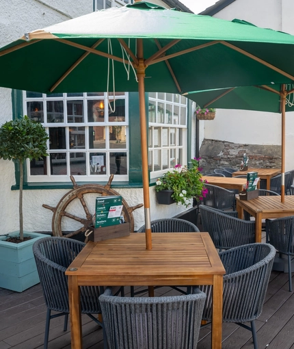 The exterior facade and seating area at the front of The Ship; with shade umbrellas on some of the wooden tables, and flower baskets hanging on the white walls of the pub.