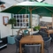 The exterior facade and seating area at the front of The Ship; with shade umbrellas on some of the wooden tables, and flower baskets hanging on the white walls of the pub.