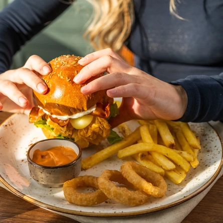 A close up view of a Crispy Chicken Burger served on a plate with fries, onion rings, and a small pot of dipping sauce. The plate sits on a cloth napkin on a wooden table, along with cutlery and a serviette, and a person sitting at the table is picking up the burger with both hands.