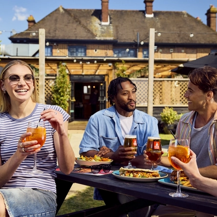 A group of friends all sat round a table, two are holding Aperol and two are holding pints of Madri in the exterior beer garden at a Flaming Grill venue.
