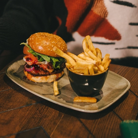 A festive decorated wooden table holding a plated burger and fries.  There is the torso of a woman holding a glass of sparkling wine sat at the table.