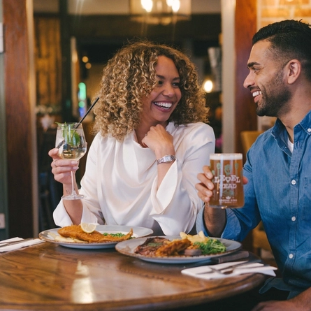 Two people sitting side by side at a small restaurant table, with plates of food in front of them. One person is holding a cocktail, and the other is holding a glass of Level Head IPA.