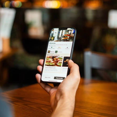 An over-the-shoulder view of a person at a wooden table holding a smartphone in one hand. The phone screen shows a venue page on the Greene King app.