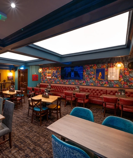 An upper floor interior restaurant seating area at the Old School House, with a long, upholstered booth seat along one side of the room, framed artwork and a TV on the wall above it, and large, square sky lights in the ceiling.