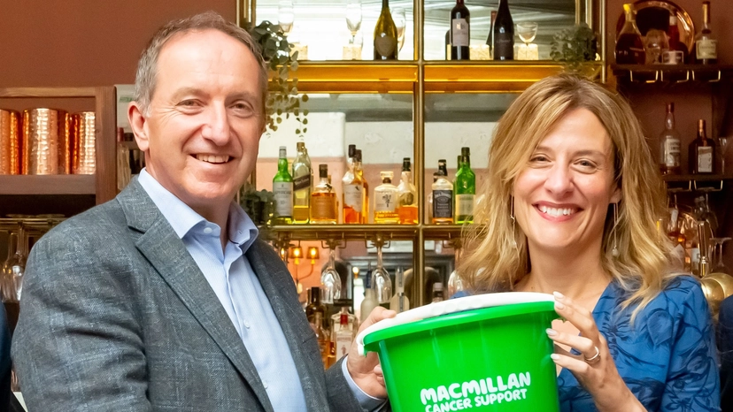 Nick Mackenzie and Gemma Peters standing in front of a bar in a pub, holding a bright green Macmillan fundraising bucket and smiling at the camera.