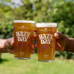 Two hands, each holding a pint of Hazy Day in the air, against a backdrop of greenery in a beer garden.