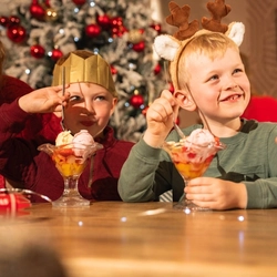 Two children sit at a festive dining table enjoying ice cream sundaes topped with fruit and cream. One child wears a reindeer antler headband and the other wears a gold paper crown from a Christmas cracker. A snow-covered Christmas tree decorated with red and gold baubles stands in the background, while drinks and festive table decorations create a warm family Christmas dining scene.