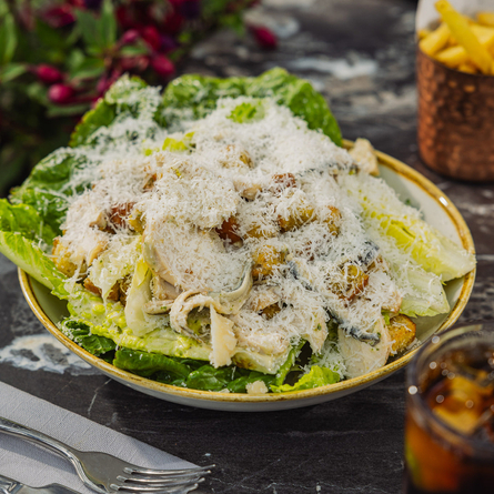 A bowled Chicken Caesar Salad dish with chips sat on a table within the exterior beer garden seating area at a Crafted venue.