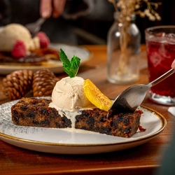 A close-up of a dessert plate featuring a slice of fruitcake topped with a scoop of vanilla ice cream, garnished with a mint leaf and an orange slice. A hand is holding a spoon, ready to take a bite.