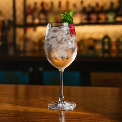 A close up view of a Blushing Hugo Spritz cocktail, served in a wine glass with ice, mint, and a dried raspberry. The glass stands on the bar inside a pub.