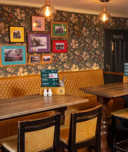 An interior restaurant seating area at The Royal Oak; with an upholstered booth seat along one side of the room, and framed artwork on the floral wallpapered wall above it.