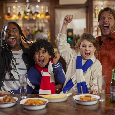 Two adults and two children sitting at a wooden table, cheering with their arms raised. Drinks and small bowls of food sit on the table in front of them.