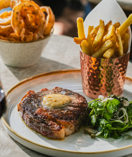 An advertising image showing a plated 35 Day Dry Aged 10oz Rib Eye Steak dish and glass of red wine sat on a table within the exterior beer garden seating area at a Crafted venue.
