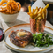 An advertising image showing a plated 35 Day Dry Aged 10oz Rib Eye Steak dish and glass of red wine sat on a table within the exterior beer garden seating area at a Crafted venue.