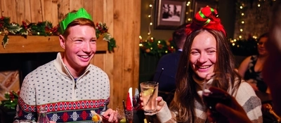 A group of people are gathered around a table enjoying a festive meal. They are wearing paper crowns, and the table is set with plates of food. The background is decorated with Christmas decorations.