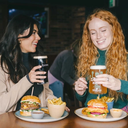 A lifestyle image of 2 friends sat within the interior restaurant seating area enjoying Burger dishes and various drinks available at Urban Core venues.