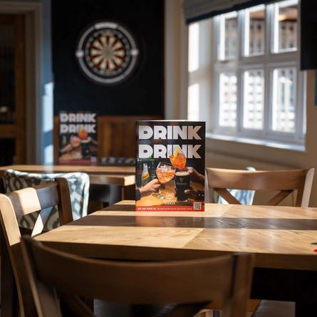 A close up view of wooden tables and chairs in the sunlit interior restaurant seating area of The Weathervane, with a dartboard visible in the background.