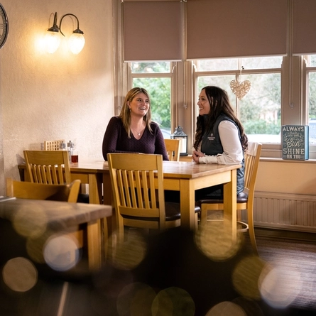 Two women sat at a table facing each and smiling.
