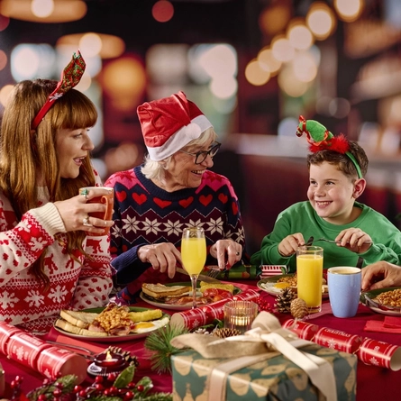 Three adults and a child sitting at a restaurant table with plates of cooked breakfast in front of them. Two glasses of juice and a mug of coffee also stand on the table along with Christmas crackers and Christmas presents; one person is holding another mug of tea or coffee; and a Christmas tree is visible in the background.