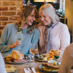 Four people sitting at a restaurant table with plates of food in front of them. One person is holding a cocktail, and a glass of wine also sits on the table.