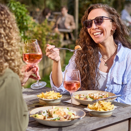 A banner showing 2 people sat at a table enjoying mains dishes with sides and glasses of rose wine within the exterior beer garden seating area at a Chef & Brewer venue.