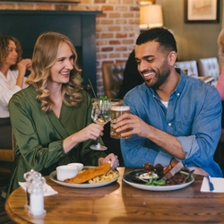 A man and woman seated at an indoor wooden table with plated food in front of them.  They are clinking their glasses together.