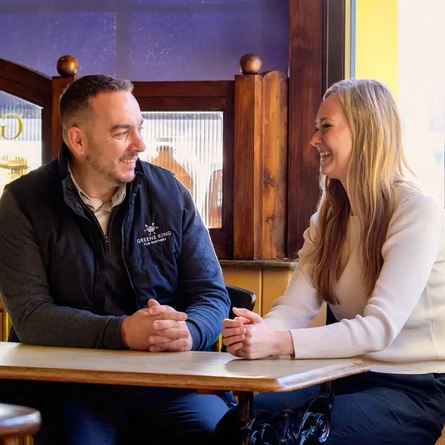 A Woman and Man sat at a table looking at each other smiling.