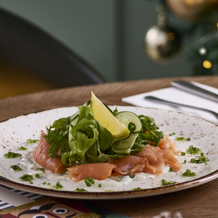 An advertising image showing a Christmas Smoked Salmon Salad Starter dish and a glass of sparkling white wine from the Festive Spread menu sat on a table within the interior restaurant seating area at a Chef & Brewer venue.