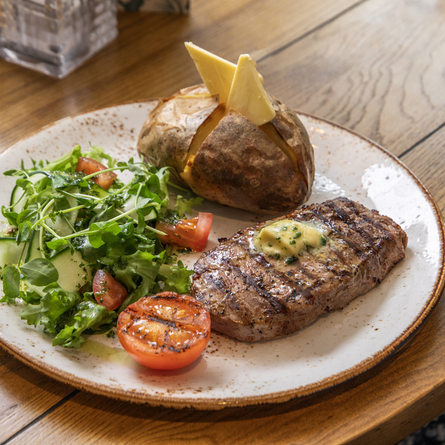 A Non-Gluten Containing Ingredients steak meal, featuring sirloin steak, a jacket potato, salad and grilled tomato, served on a plate which sits on a wooden table.