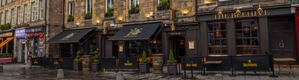 The exterior facade, signage, and seating area of The Beehive Inn in Edinburgh, with potted topiary trees, awnings hanging over the tables and chairs, and flower boxes on the upper windowsills.