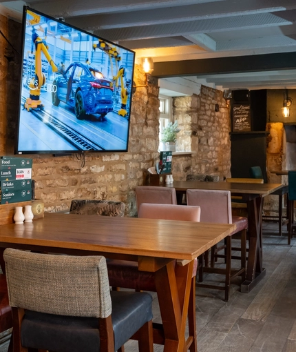 An interior restaurant seating area at The Royal Oak; with wooden flooring, wooden beams on the ceiling, and a large TV on a stone wall.