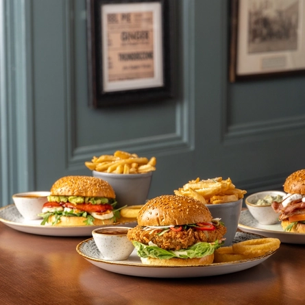Three plates of burgers, chips, onion rings and sauce, sitting on a wooden table.