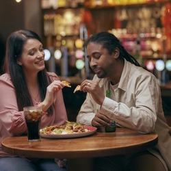 A man and woman sitting at a table with a bottle of Heineken and a glass of Coca Cola, sharing a plate of nachos.