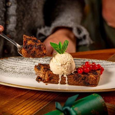 A wooden table, decorated with festive decoration is  holding a plated Christmas Pudding with ice cream.  A person seated at the table is holding a spoon with some of teh Christmas Pudding on it.  Only the torso is visible in the image.